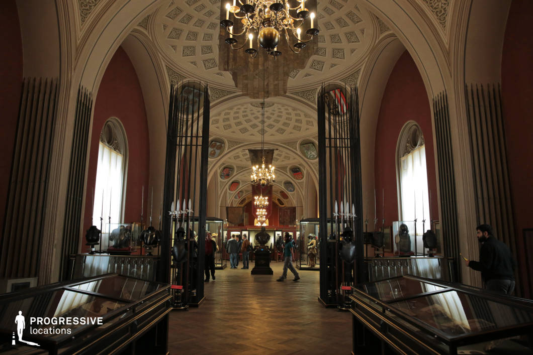 This stately exhibition hall, dedicated to military history, features soaring arched ceilings adorned with intricate geometric patterns and heraldic emblems. The deep red walls and dim, atmospheric lighting create a sense of solemnity and reverence. Display cases lined with polished glass showcase historical armor, weaponry, and artifacts, while tall wrought-iron gates with spear-like details add to the hall’s regal and fortified aesthetic. Chandeliers cast a warm glow, accentuating the structured symmetry of the space. This location is ideal for filming historical documentaries, medieval or military-themed dramas, or museum-set intrigue, offering a setting rich in heritage and authority.
