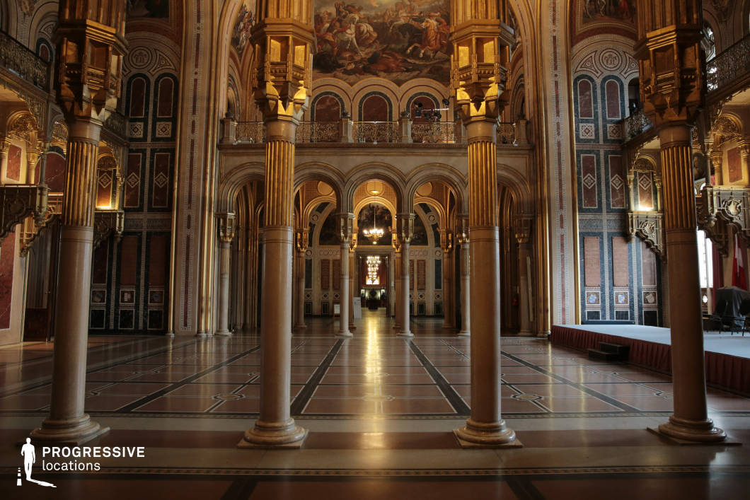 This majestic hall exudes a sense of grandeur with its towering fluted columns, gilded capitals, and richly decorated walls featuring intricate geometric patterns and classical motifs. The symmetrical arrangement of arched doorways and balconies enhances the depth of the space, while the polished floor reflects the ambient lighting, creating an almost ethereal atmosphere. Large murals depicting historical and mythological scenes add to the sense of opulence and narrative depth. This location is ideal for historical epics, fantasy productions, or regal ceremonies, offering a setting that conveys power, tradition, and timeless elegance.