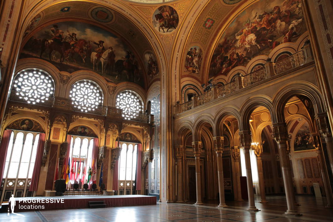 This grand hall exudes historical significance with its soaring arched ceilings adorned with intricate battle-themed frescoes and gold detailing. Expansive stained-glass windows with elaborate rose patterns cast diffused light onto the richly decorated walls, creating a dramatic yet refined ambiance. A series of stately columns and elegant archways frame the space, adding depth and symmetry to the composition. The combination of regal ornamentation, classical artistry, and an elevated gallery makes this location ideal for filming period dramas, political gatherings, or ceremonial events, offering a setting that conveys power, history, and prestige.