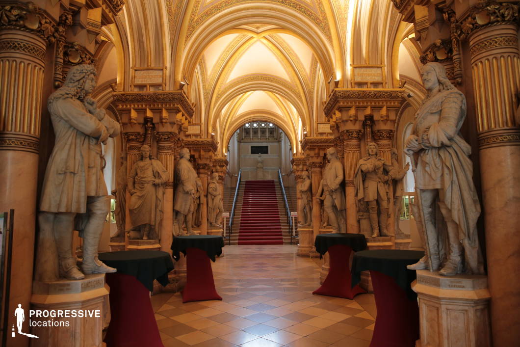 This grand hall exudes historical prestige, featuring towering marble statues of distinguished figures lining both sides of a regal red-carpeted staircase. The high, pointed arches adorned with intricate gold detailing create a cathedral-like ambiance, enhanced by the symmetry of the space. Soft, warm lighting accentuates the rich architectural textures, from the ornately carved columns to the patterned tiled floor. The interplay of grandeur and solemnity makes this an ideal setting for historical dramas, royal court scenes, or epic period films, providing a visually striking backdrop for moments of power, legacy, or intrigue.