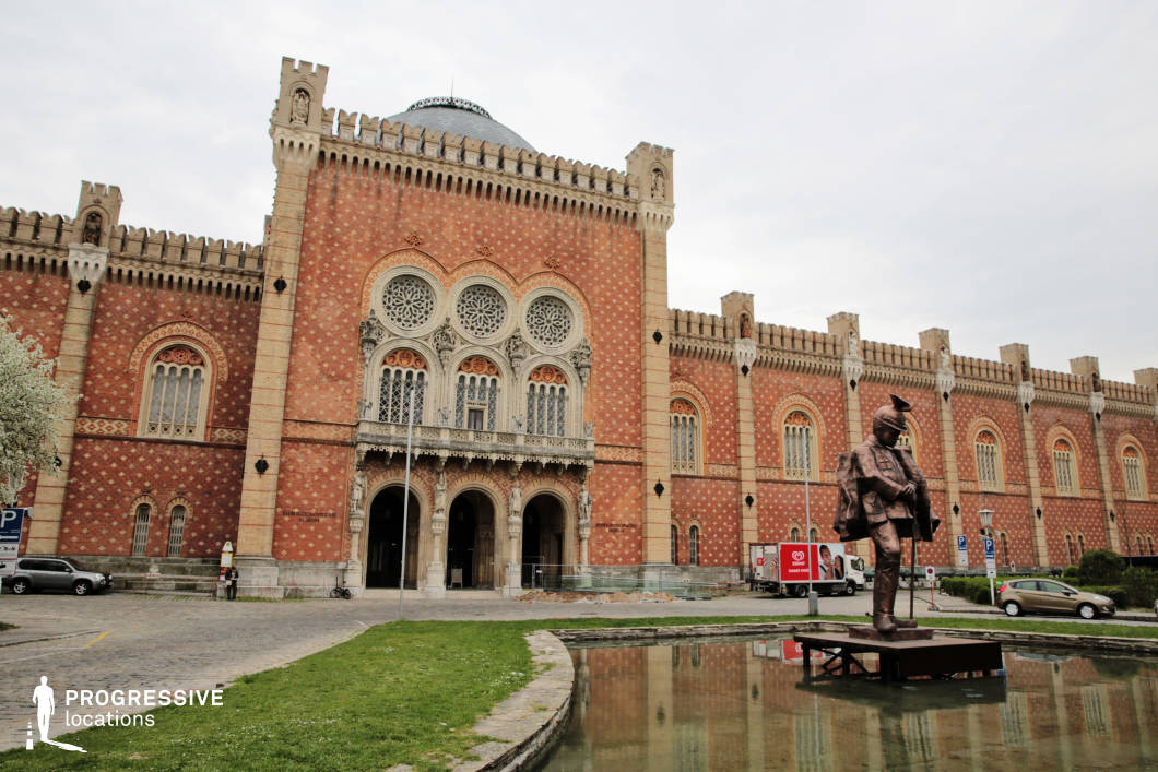 This imposing Neo-Gothic and Moorish Revival-style building features an intricate red-brick facade adorned with decorative patterns, arched windows, and elaborate stone tracery. The central entrance is framed by three grand archways, topped by a series of ornate circular rose windows, while crenelated towers add a fortified, historic character. A bronze statue stands before a reflective water feature, reinforcing the site's military significance. This location is ideal for historical documentaries, period dramas, or productions requiring a majestic and institutional backdrop, offering a blend of architectural grandeur and a sense of historical depth.