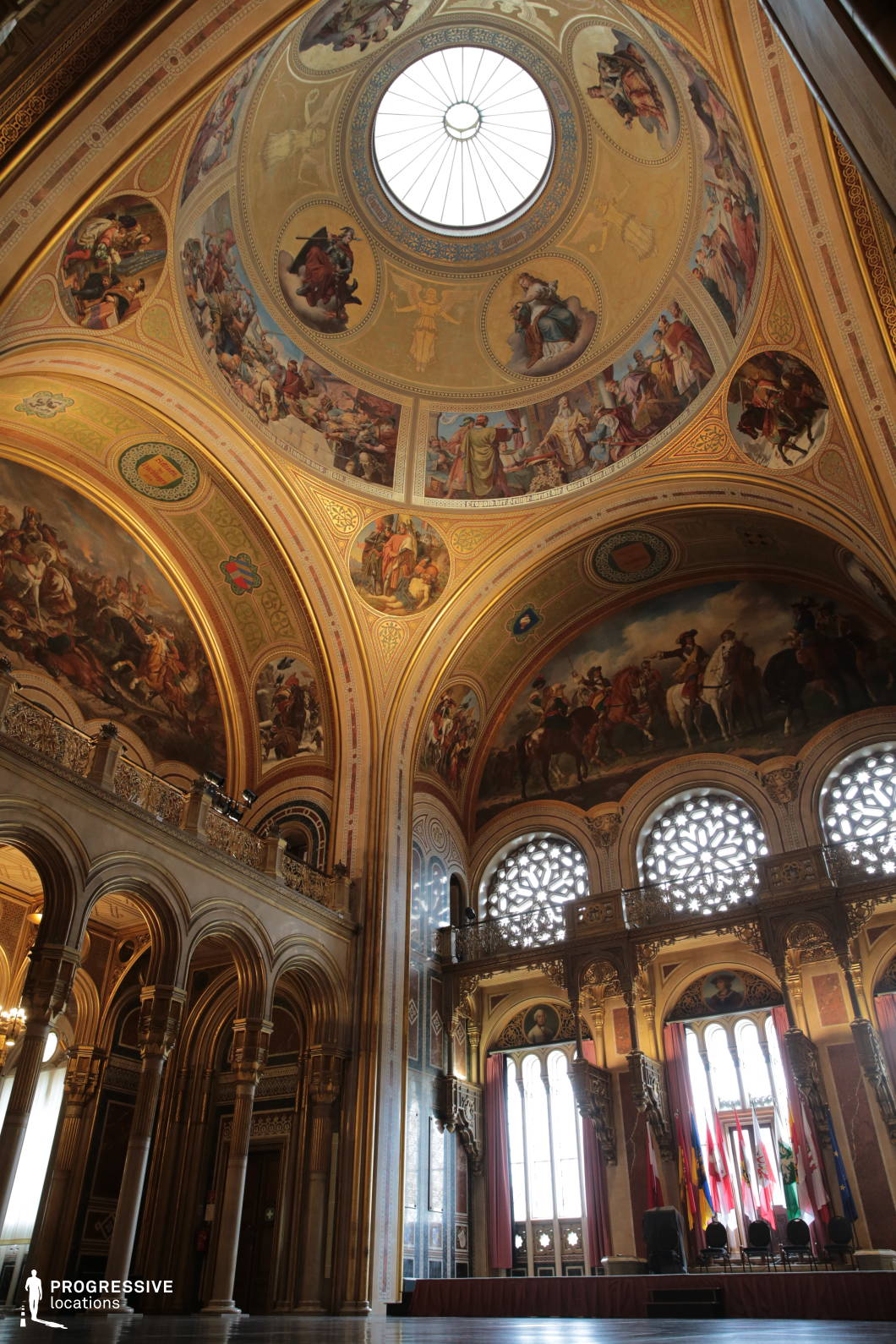 This grand hall exudes historical significance and architectural splendor, featuring an intricately decorated domed ceiling adorned with elaborate frescoes depicting historical and mythological scenes. The richly gilded arches, detailed friezes, and sculpted ornamentation create a regal ambiance, complemented by towering arched windows with delicate tracery that filter in natural light. A grand balcony with decorative railings adds depth and scale to the space, while national flags enhance its formal and institutional character. This location is ideal for filming historical epics, political dramas, or period pieces requiring a setting of authority, grandeur, and cultural heritage.