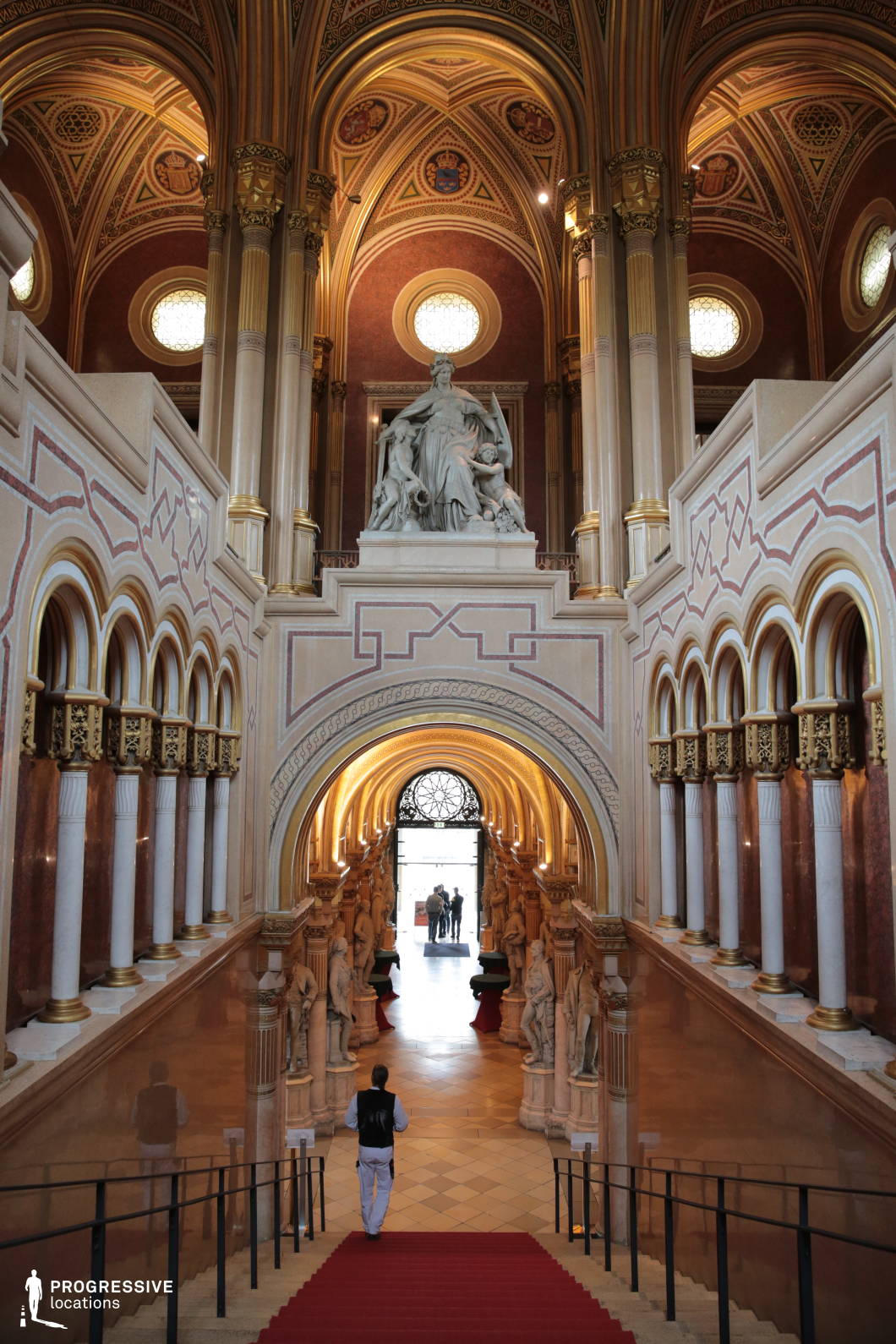 This grand staircase exudes Neo-Gothic splendor, featuring soaring vaulted ceilings adorned with intricate geometric and heraldic patterns. The richly decorated space is framed by a series of arches, gilded columns, and marble-clad walls, leading the eye toward a central statue seated above an ornate archway. Rows of classical sculptures line the lower hall, enhancing the sense of historical reverence. The warm color palette of gold, ochre, and deep red, along with the regal red carpet, contributes to the imposing yet refined atmosphere. This location is ideal for historical epics, fantasy films, and regal dramas, providing an authoritative and visually striking backdrop for scenes of power, heritage, or ceremonial grandeur.