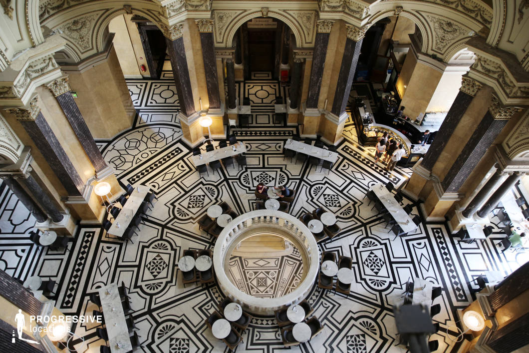 This breathtaking neoclassical interior features a striking black-and-white geometric patterned marble floor, framed by towering, intricately carved stone columns and grand archways. The circular central opening, surrounded by elegant seating, adds depth and symmetry to the composition. Soft lighting highlights the rich architectural details, while marble-topped tables and plush chairs enhance the luxurious atmosphere. The soaring ceilings, elaborate moldings, and classical design elements create an opulent setting, ideal for historical dramas, high-society period films, or refined commercial productions, evoking timeless elegance and sophistication.