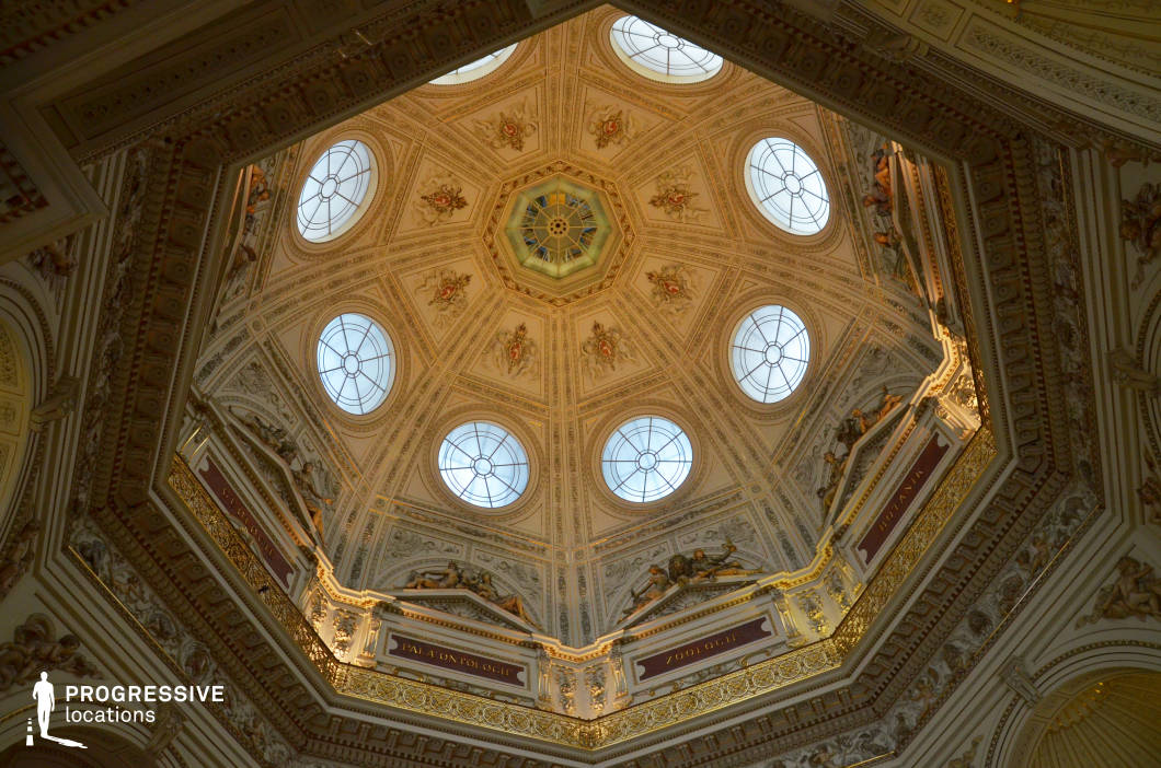 This breathtaking domed ceiling showcases exquisite neoclassical craftsmanship, featuring intricate gilded moldings, sculptural reliefs, and delicate frescoes. The symmetrical octagonal design is adorned with circular skylights, allowing soft natural light to filter through, highlighting the fine detailing of the artwork. Below the dome, elaborate architectural elements, including gold-accented inscriptions and sculpted figures, contribute to the grandeur of the space. This location is ideal for filming historical dramas, academic or museum settings, and sophisticated cultural narratives, offering an atmosphere of knowledge, prestige, and timeless elegance.