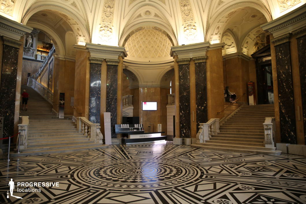 This majestic entrance hall showcases grand neoclassical architecture with towering marble columns, intricate vaulted ceilings adorned with sculptural reliefs, and a striking black-and-white patterned marble floor that serves as a visual centerpiece. Twin sweeping staircases with ornate balustrades lead to upper levels, adding to the hall’s imposing symmetry and sense of scale. The polished stone surfaces, including dark marble paneling and gilded decorative elements, enhance the opulent yet formal atmosphere. This location is ideal for filming historical dramas, intellectual thrillers, or prestigious institutional settings, offering a refined and authoritative backdrop suited for museum, palace, or high-society scenes.