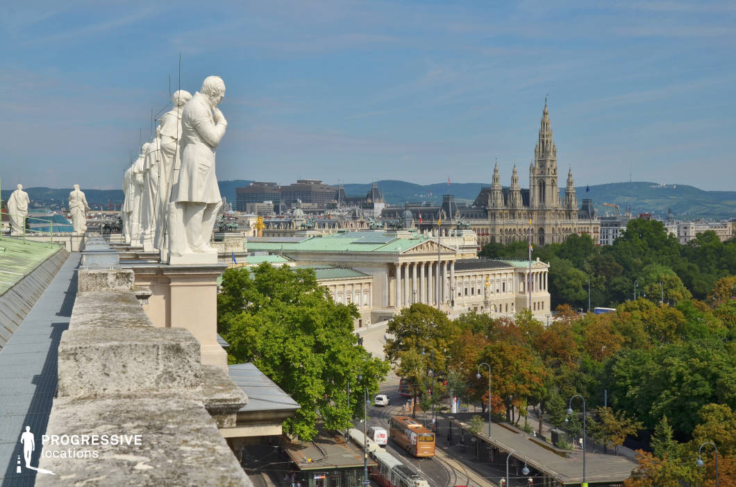 This stunning rooftop vantage point offers a panoramic view of an architecturally rich city, framed by a series of classical white statues standing atop an ornate balustrade. In the background, grand landmarks, including a neoclassical parliament building with a green copper roof and a towering Gothic-style city hall, rise above a lush expanse of tree-lined streets. The urban layout blends historic grandeur with modern elements, featuring tram lines and contemporary structures against rolling hills in the distance. This location is ideal for filming political dramas, historical narratives, or intellectual thrillers, providing a visually captivating setting with a strong European identity and a blend of old-world elegance and urban dynamism.