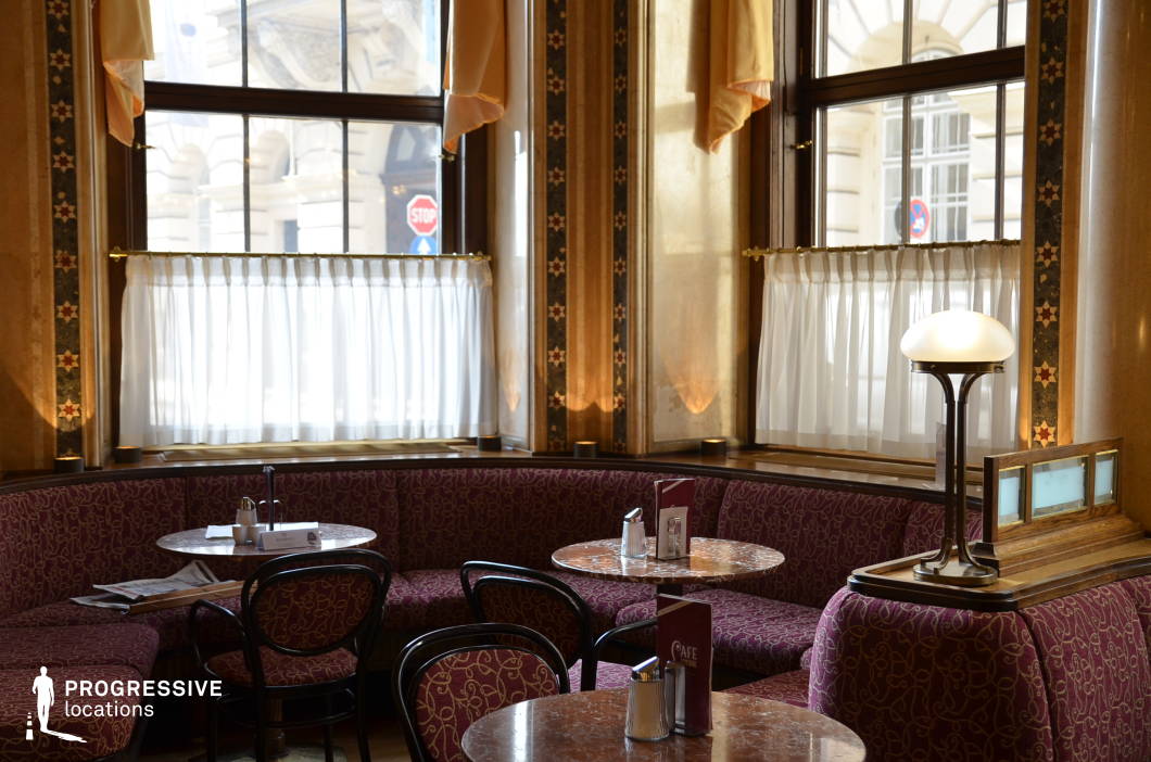 This refined interior captures the elegant atmosphere of a historic European café, featuring richly upholstered corner banquettes and small, round marble-topped tables. The space is defined by tall windows dressed with semi-sheer white curtains and framed by decorative columns adorned with geometric star motifs and warm-toned marble paneling. A frosted glass lamp with a bronze base provides soft illumination and contributes to the classic ambiance. The palette combines deep burgundy, gold, and cream tones, complemented by dark wooden furniture and detailed trim. The layout is intimate, with closely spaced seating arranged to encourage quiet conversation or reflective solitude. Outside the windows, architectural details from adjacent buildings add to the period charm. This location offers a timeless, cultivated backdrop ideal for scenes requiring an upscale, old-world café setting. It is particularly suitable for dramatic or contemplative moments, with its rich textures and historical character lending authenticity and visual depth.