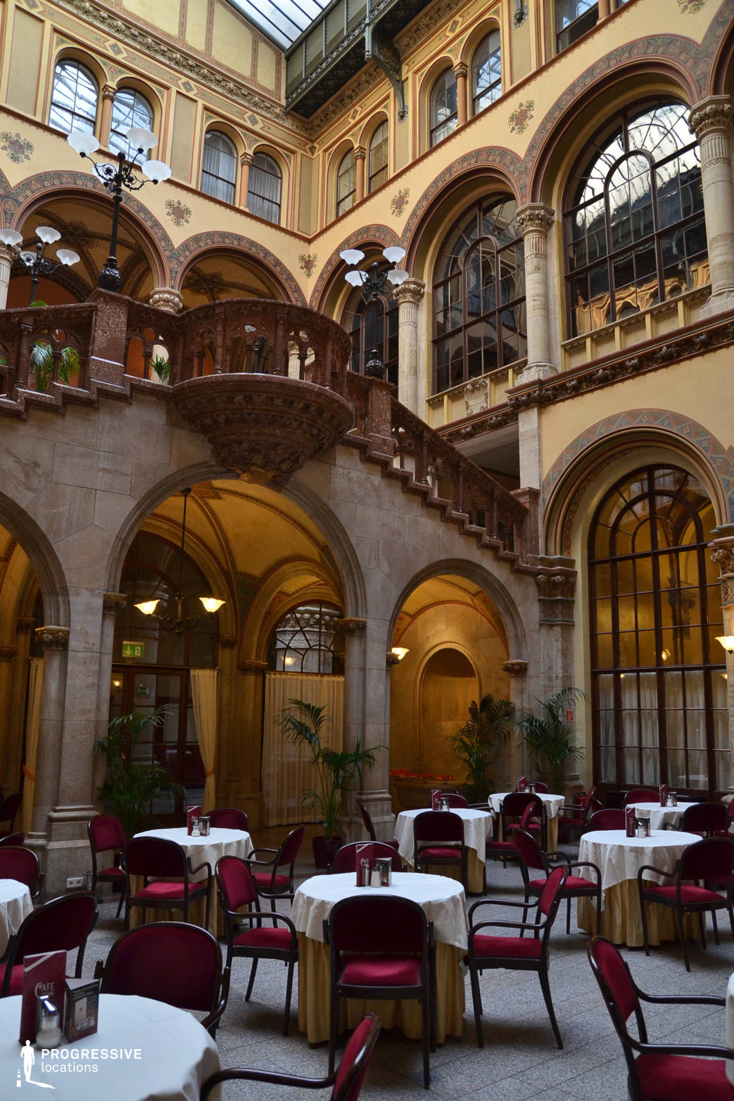 This majestic interior features a café set within a vaulted atrium of a historic building, enclosed by elaborately decorated arches and columns. The architectural style combines neo-Renaissance and Gothic Revival elements, with ornamental stucco work, floral motifs, and a finely carved stone staircase that leads to an upper balcony. Large arched windows and an overhead glass ceiling allow natural light to flood the space, enhancing its grand proportions and elegant ambiance. The café area below is arranged with round tables draped in cream-colored cloths, each paired with dark wooden chairs upholstered in deep red. Tall potted palms and antique-style lamps add a refined softness to the formal geometry of the space. This location is ideal for sophisticated period scenes, literary or cultural settings, and luxurious indoor productions. Its dramatic height, classical detailing, and well-preserved historical elements offer a cinematic atmosphere that evokes timeless elegance and European café tradition.