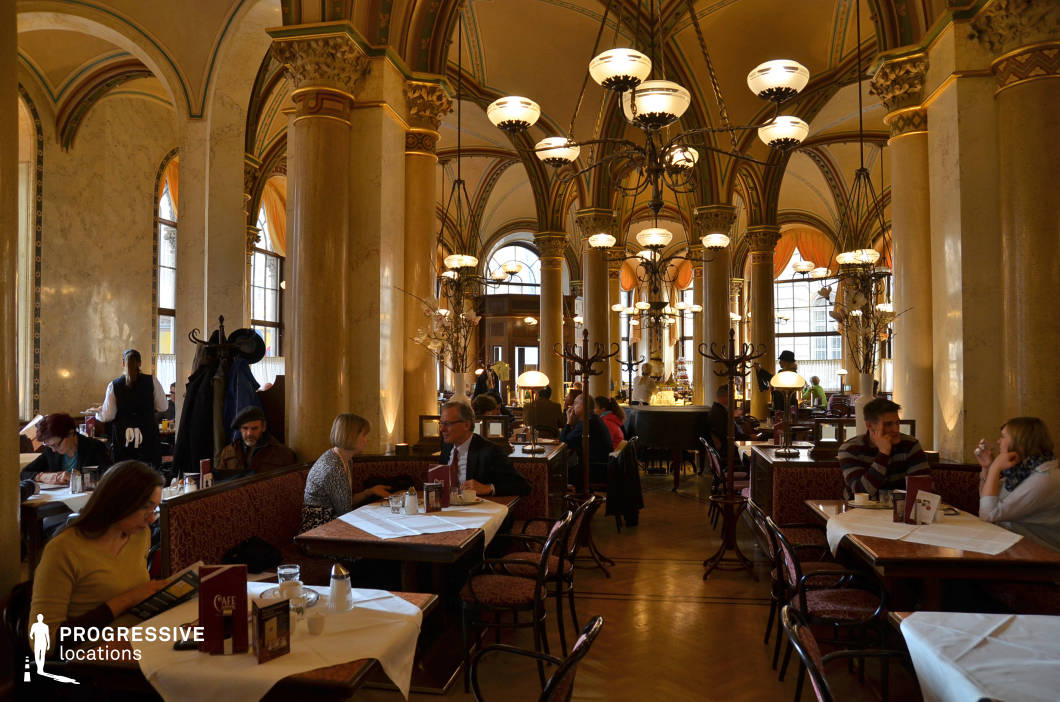 This richly decorated café hall is housed within a historic building, featuring vaulted ceilings supported by ornate marble columns with Corinthian-style capitals. The architecture is defined by symmetrical arches, ribbed vaulting, and soft golden hues enhanced by ambient lighting from large globe chandeliers and art deco lamps. Large arched windows allow filtered natural light to complement the elegant interior illumination. The seating arrangement consists of wooden tables with crisp white tablecloths and classic upholstered banquettes and chairs in burgundy tones. The parquet flooring adds warmth and continuity to the space, while decorative coatracks and floral accents provide functional charm. The central bar area in the background offers a focal point and depth to the composition. This location is ideally suited for refined, culturally rich scenes requiring historical grandeur or a distinguished social setting. Its detailed architecture and carefully composed lighting support both period and contemporary narratives with sophistication and visual texture.