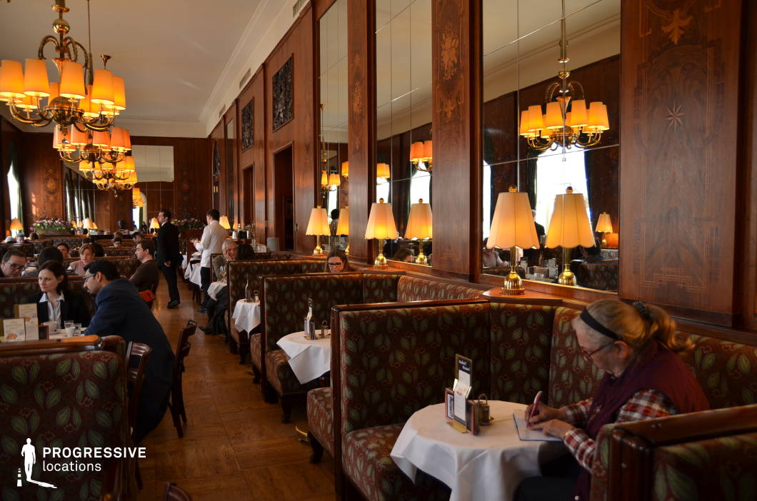 This elegant café interior showcases a timeless design defined by high-backed banquettes upholstered in patterned fabric and separated by polished wood dividers. The walls are clad in intricately inlaid wood paneling with floral motifs and integrated mirrored sections that enhance spatial depth. Each seating alcove is softly lit by brass-based table lamps with cream-colored shades, creating a warm and intimate atmosphere. The central aisle is flanked by chandeliers suspended from a high ceiling, casting a golden glow that complements the natural tones of the flooring and furnishings. The layout supports both quiet solitude and sociable interaction, with white-clothed tables uniformly set for service. This location is ideal for film scenes or photo shoots that require a refined, traditional European café setting. It provides a sophisticated backdrop for dialogue, reflection, or cultural ambiance, combining historic charm with structured visual symmetry.