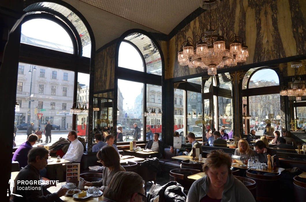 This refined urban café interior combines historic grandeur with lively street-facing openness. Defined by tall, arched windows and mirror-clad walls, the space draws in abundant natural light, creating visual continuity between the café and the classic European cityscape beyond. Crystal chandeliers hang from the vaulted ceiling, contributing to the upscale ambiance alongside dark leather seating and polished wooden tables. The interior features a rich material palette, including dark veined marble walls and brass accents, while the mirrored surfaces visually extend the space and reflect the bustling street scenes. Outside, trams and pedestrians animate the city backdrop, reinforcing the café’s position within a vibrant metropolitan setting. Ideal for productions seeking a cultured, metropolitan atmosphere with architectural sophistication, this location blends vintage elegance with everyday accessibility, making it suitable for dramatic exchanges, reflective moments, or social interactions set against a European urban café backdrop.