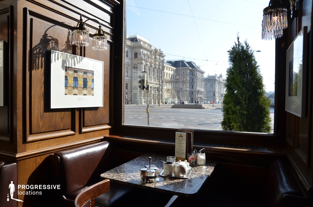 This image captures a cozy corner table in a traditional European café, offering an unobstructed view through a large window onto a grand urban streetscape. The table, finished with a polished granite surface, is neatly set with a white teapot, glasses, sugar containers, and a menu, suggesting an inviting space for quiet conversation or solitary reflection. The setting is framed by dark wood paneling and leather-upholstered seating, contributing to the overall warmth and elegance. Crystal-accented sconces cast a delicate light over framed artwork on the walls, reinforcing the refined interior styling. The view outside reveals historic buildings with classical facades, adding a cinematic quality to the location. This corner is ideal for intimate café scenes, interviews, or contemplative moments, offering a harmonious blend of interior richness and exterior urban beauty. Its strategic composition and natural lighting make it particularly suitable for productions requiring authenticity, refinement, and visual storytelling through framing.