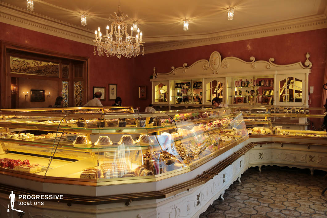 This richly decorated interior showcases a grand pastry counter set within an elegant, old-world café. The counter, finished in cream and gold detailing, is filled with meticulously arranged pastries, cakes, and confections under soft-glow glass displays. The decor features baroque-inspired cabinetry and curved wooden elements, contributing to a sense of refined tradition. The room is painted in warm terracotta tones and enhanced by a crystal chandelier that adds a touch of luxury and soft overhead lighting. Wall-mounted display shelves hold boxed confections and decorative items, reinforcing the artisanal and heritage-driven ambiance. With its vintage charm and visual richness, this space is ideal for commercial or period film settings involving culinary artistry, nostalgic elegance, or café culture. The abundance of reflective surfaces and ornate architectural features makes it highly photogenic and adaptable for a variety of stylistic directions. 