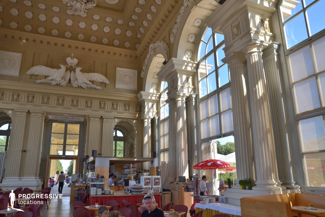 This location features an impressive café set within a grand neoclassical hall, marked by tall, fluted columns, ornate entablatures, and vast arched windows that flood the space with natural light. The soaring ceiling is richly decorated with coffered panels and a dramatic sculptural relief of double-headed eagles, evoking imperial grandeur. The interior includes a mix of traditional and modern elements—simple wooden service counters contrast with the historical architecture, while contemporary café seating introduces a relaxed, functional element. The windows are equipped with large blinds, which soften the daylight and create dynamic shadow play across the space. This site offers an exceptional backdrop for high-end commercial shoots, historical dramas, or upscale lifestyle content. Its combination of monumental scale, classical elegance, and accessible modern furnishing makes it highly versatile for various creative applications.