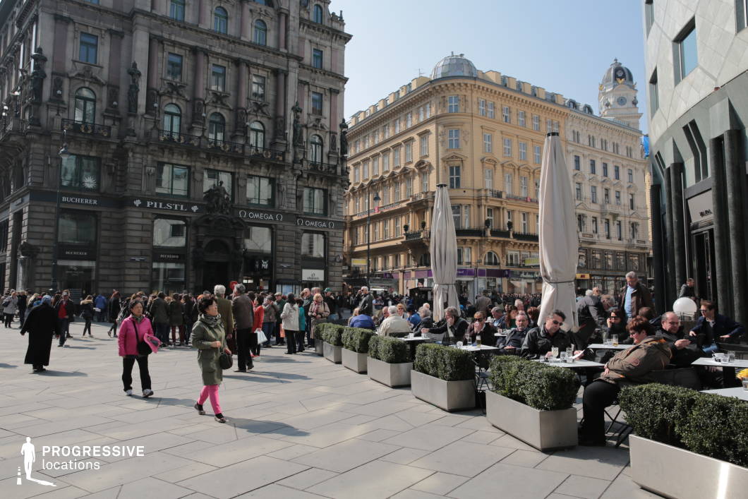 This urban location features a vibrant café terrace nestled within a grand European square, surrounded by richly ornamented historic architecture. The outdoor seating area is lined with minimalist metal tables and chairs, complemented by large planter boxes and closed parasols, offering a clean contrast to the detailed façades of the surrounding buildings. The square itself is paved with light stone tiles, forming a visually cohesive pedestrian space that supports high foot traffic and creates a lively, bustling atmosphere. The mix of Renaissance Revival and early modern architecture provides an elegant backdrop, ideal for portraying upscale city life, social scenes, or cosmopolitan settings. This location is perfectly suited for lifestyle shoots, fashion editorials, or commercial productions that require a dynamic urban setting with architectural richness and a classic European ambiance.
