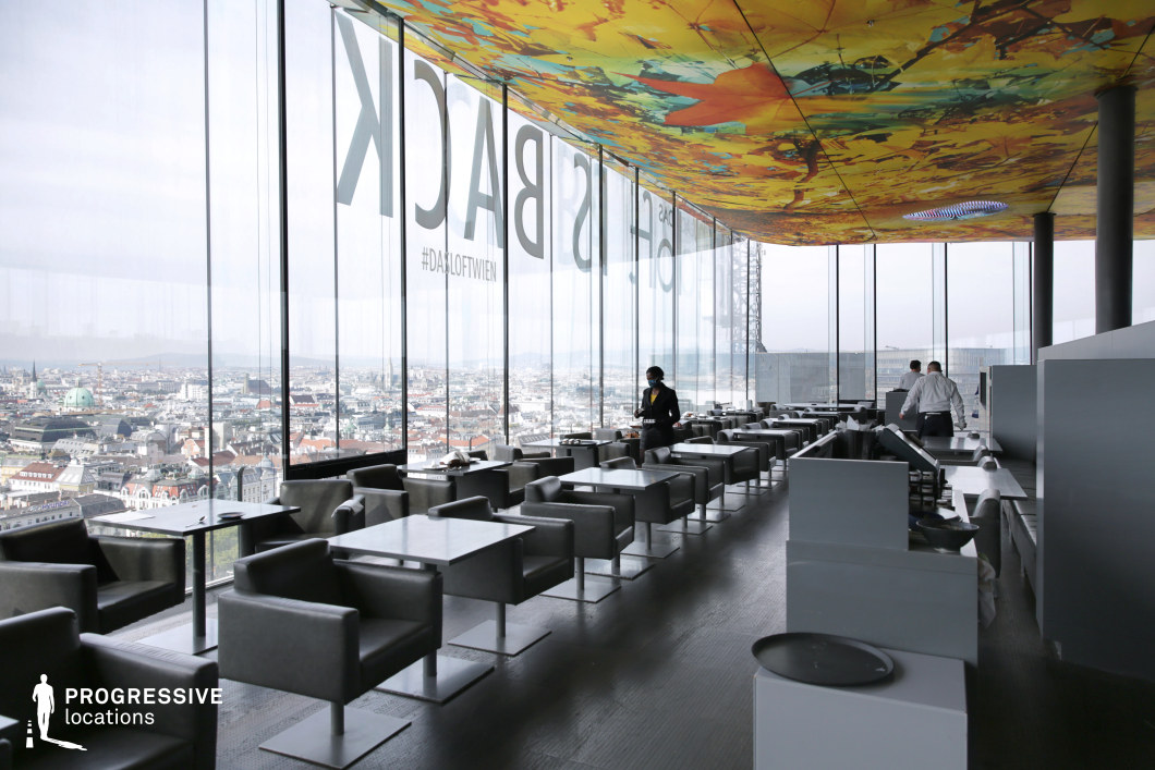 This elevated rooftop lounge boasts floor-to-ceiling glass walls offering a sweeping panoramic view over a densely built European cityscape. The sleek, minimalist interior is furnished with matte grey square tables and plush leather armchairs, arranged in neat rows to maintain clear lines and visual order. A standout feature is the bold, abstract ceiling mural in vivid orange and yellow tones, adding a dramatic artistic contrast to the otherwise monochromatic design. The space exudes a contemporary, upscale atmosphere, ideal for elegant dining scenes, luxury product placements, or high-end commercial shoots. The expansive glazing not only floods the interior with natural light but also frames the skyline as a dynamic, ever-changing backdrop, enhancing the location’s visual depth and cinematic potential.
