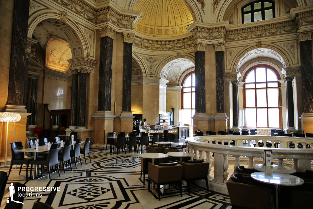This opulent café is set within a historic museum’s grand rotunda, combining monumental classical architecture with modern seating. Towering marble columns, intricately decorated arches, and a gilded coffered dome ceiling frame the space, while large arched windows flood the hall with natural light. The black-and-white patterned marble flooring adds visual rhythm and a touch of regal elegance. Contemporary furnishings—including black chairs, polished tabletops, and a circular balustrade separating seating zones—create a refined contrast within the palatial setting. Ideal for high-end fashion shoots, historical or intellectual-themed productions, or elegant dining scenes, this location offers a striking balance of sophistication, heritage, and architectural drama.