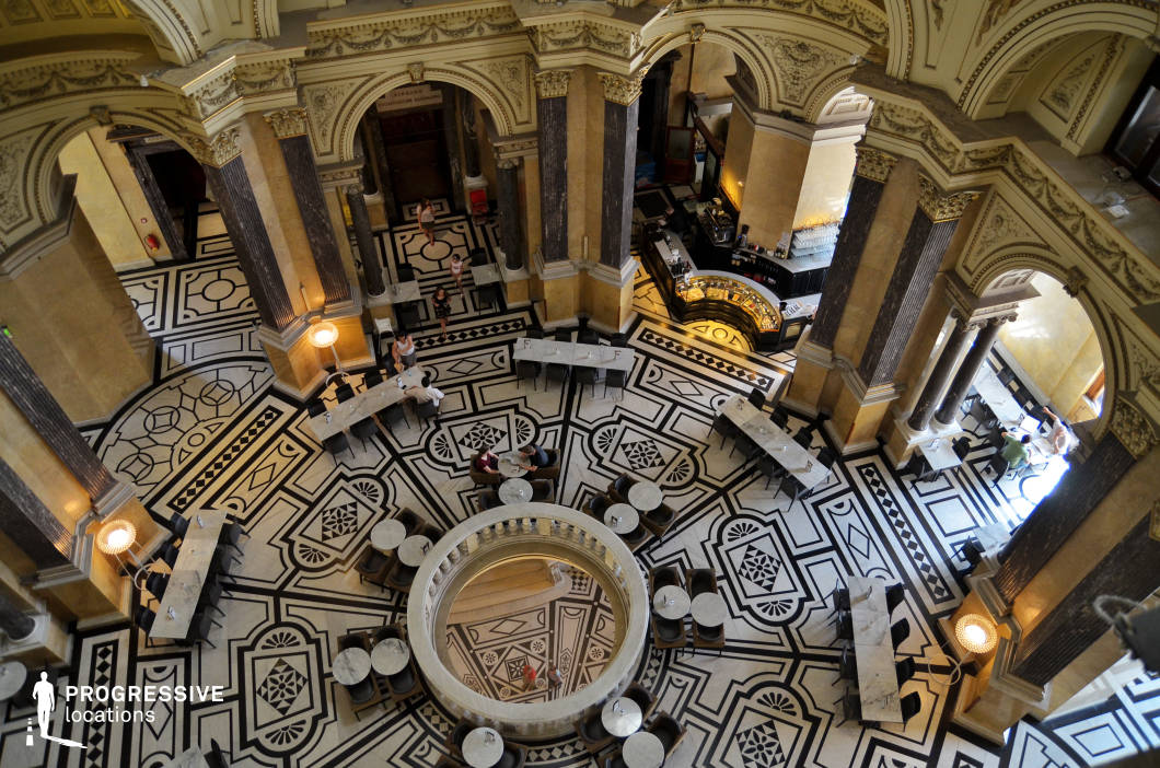 This top-down view reveals the dramatic geometry and elegance of a grand museum café set within a historic rotunda. The black-and-white mosaic floor showcases intricate patterns in perfect symmetry, encircling a central balustraded opening to a lower level. High arched doorways, classical columns, and ornamental cornices reinforce the location’s neo-baroque architectural richness. Elegant marble-topped tables and contemporary black chairs are arranged in a structured layout that balances modern café culture with heritage ambiance. The overall visual effect is one of refined formality and artistic grandeur, making this location ideal for stylized fashion shoots, formal cinematic scenes, or settings evoking cultural sophistication.
