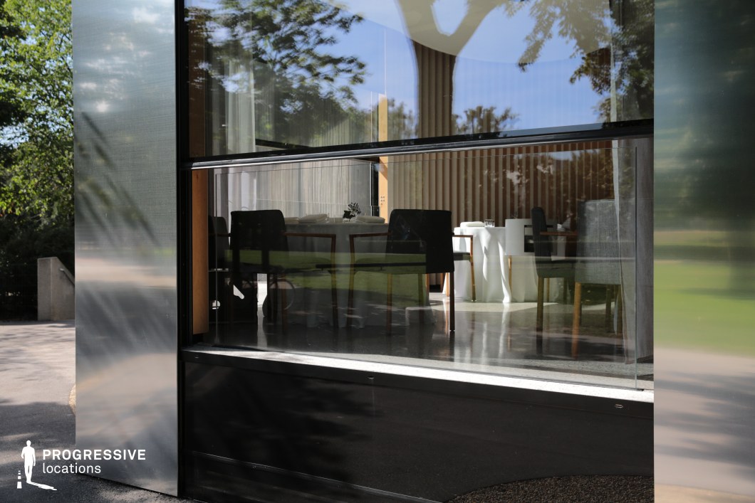 This contemporary restaurant façade showcases a seamless fusion of brushed metal surfaces and expansive floor-to-ceiling glass panels, offering a reflective view into an elegant dining area. The interior features minimalist decor with clean lines, light wood slats, and monochromatic furniture including dark upholstered chairs and crisp white table settings. The architectural design emphasizes transparency and simplicity, creating a visual dialogue between the refined indoor space and the surrounding greenery. Ideal for modern lifestyle productions, upscale culinary scenes, or sleek urban design-focused shoots, this location lends itself to sophisticated and understated visual storytelling.