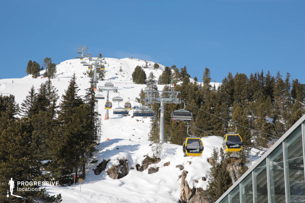 This high-altitude location features a dual transportation infrastructure composed of a modern ski lift and enclosed yellow cable cars, both running parallel up a snow-covered mountainside. The slope is densely forested with tall, dark green conifers that provide strong vertical accents and contrast with the crisp white snow. The terrain rises in a gradual incline toward a rounded summit, with rocky outcrops partially exposed among the snowbanks. The dominant materials include steel for the lift structures and glass for the nearby angular-roofed station visible in the foreground, contributing to a contemporary, utilitarian alpine aesthetic. The color palette is defined by stark contrasts between white snow, green trees, and the bold yellow of the gondolas. The open composition offers a visually expansive setting with clear depth, ideal for wide-angle shots or panoramic filming. This location’s infrastructure and natural elements make it suitable for showcasing dynamic mountain activity, winter sports environments, or remote alpine settings with a functional, structured layout.