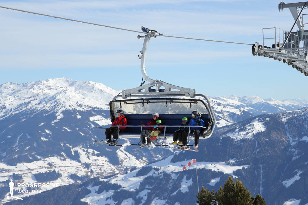 This location captures a high-altitude open chairlift system suspended above a vast alpine landscape. The foreground is dominated by the metallic structure of the chairlift, featuring a sleek, curved frame and deep blue cushioned seats, indicating a modern and functional design. The background reveals a panoramic view of snow-covered mountains and densely forested valleys, forming a layered topography that recedes into the distance. The horizon is composed of rounded peaks, offering a dramatic yet serene natural backdrop. The materials primarily include brushed metal and synthetic padding, with the chairlift's geometric form emphasizing industrial precision against the organic curves of the surrounding terrain. The perspective is slightly tilted upward, enhancing the sense of elevation and spatial openness. The composition is ideal for showcasing verticality and vastness, making this setting particularly suitable for productions requiring expansive, visually impactful mountain scenery.