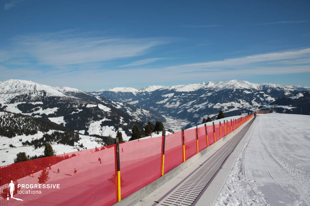 This elevated mountain location showcases a wide panoramic view of snow-covered alpine ridges, valleys, and forested slopes. A prominent element in the composition is the linear travelator system running alongside a bright red safety net, guiding the viewer’s eye deep into the mountainous landscape. The mechanical path appears to serve a transport or beginner lift function, providing structured access along the ridge. The terrain features a combination of undulating forested hills and expansive snowfields, with deep valleys carving through the midground and distant peaks creating a layered, immersive backdrop. The color palette contrasts clean whites with dark evergreen tree clusters and the bold red safety fencing, adding rhythm and boundary to the natural environment. This location is ideal for showcasing large-scale alpine settings with clear geometric orientation and expansive views, making it well-suited for productions emphasizing high-altitude perspective, environmental scale, or structured movement through rugged terrain.
