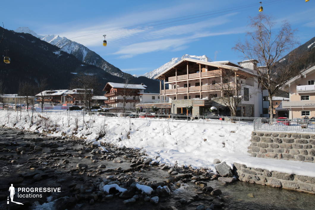 This serene alpine townscape is centered around a gently flowing mountain stream bordered by snow-covered embankments and a low stone retaining wall. The composition includes a variety of chalet-style and modern buildings with wooden balconies and pitched roofs, blending traditional architectural forms with contemporary design. In the background, the scene is framed by densely forested mountain slopes and distant snowy peaks, reinforcing the town’s embeddedness in its alpine setting. A cable car line with suspended yellow gondolas traverses the upper part of the image, adding vertical dynamism and suggesting year-round accessibility to higher elevations. The visual balance between natural and built environments is reinforced by the stream's organic flow and the structured alignment of the buildings and streetscape. This location provides a versatile setting for productions that require a small-town alpine atmosphere with scenic views, accessible infrastructure, and strong integration between urban and natural elements.