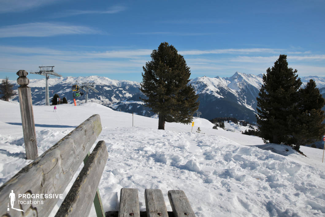 This tranquil mountain location features a weathered wooden bench positioned on a snowy slope, offering a natural viewpoint toward a dramatic alpine panorama. The foreground is defined by rough, uneven snow cover and the rustic texture of the bench, which adds a grounded human element to the vastness of the surrounding environment. A few evergreen trees are scattered across the hillside, their dark green foliage contrasting sharply with the white snow. In the midground, a cable car system with yellow gondolas traverses the slope, supported by minimalistic steel pylons that introduce vertical and geometric elements. The mountainous backdrop includes multiple rugged peaks and ridges, creating a layered composition that emphasizes scale and depth. This setting is ideal for capturing moments of stillness and reflection within a powerful alpine context. The interplay between natural materials and ski infrastructure offers versatility for both atmospheric and practical mountain scenes.