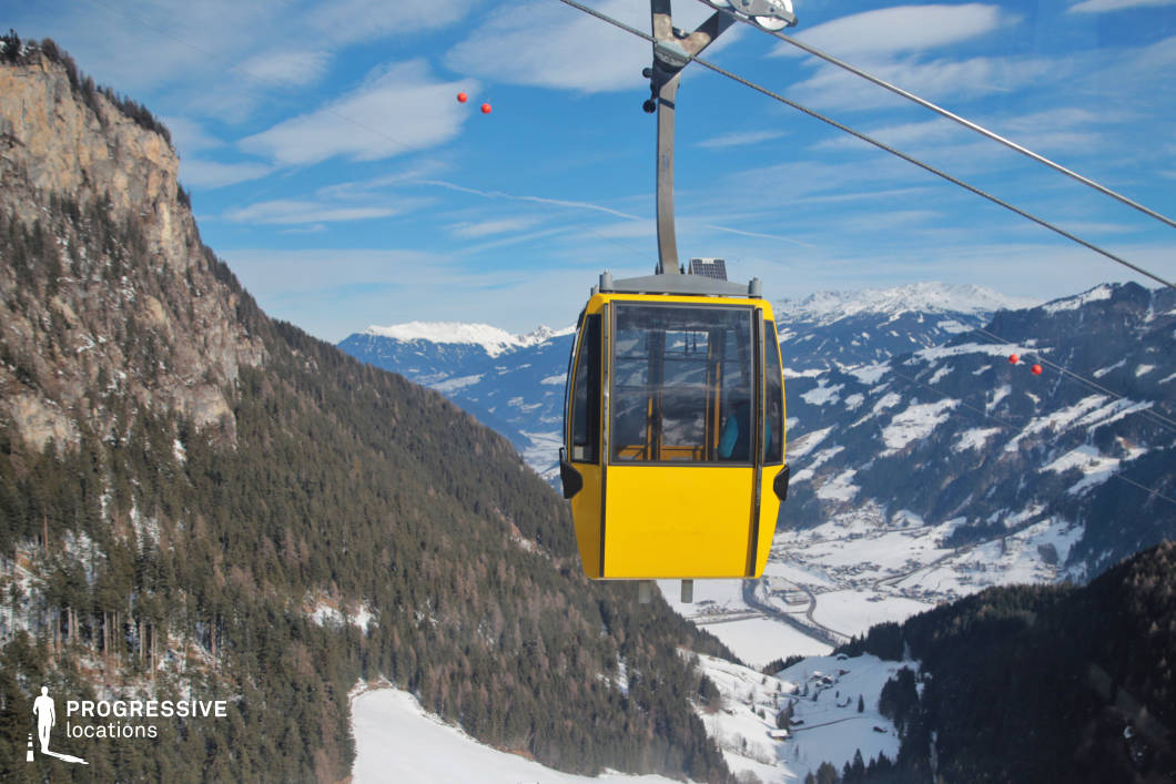 This striking aerial scene centers on a bright yellow gondola suspended from a cableway system, captured mid-journey over a vast alpine valley. The gondola is framed against a dynamic backdrop of steep forested slopes and snow-covered terrain, which descend into a patchwork of meadows, forests, and mountain villages far below. The cable lines stretch diagonally across the image, adding a sense of movement and structural rhythm. The surrounding landscape includes dramatic geological formations such as exposed rock faces and densely wooded ridgelines, contributing to the visual contrast between natural ruggedness and engineered precision. The gondola itself, composed of glass and metal, serves as a functional focal point and a visual anchor within the expansive setting. This location is highly suitable for dynamic aerial or transitional sequences in film or advertising, emphasizing elevation, vertical scale, and the immersive experience of high-altitude transport through scenic alpine terrain.