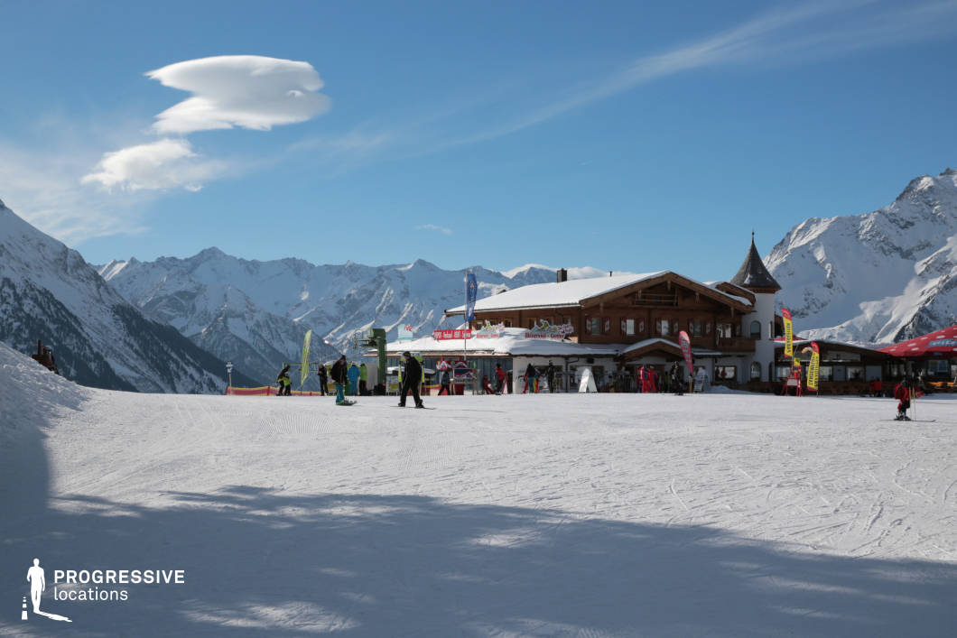 This high-altitude scene features a traditional alpine lodge situated on a wide, groomed plateau with a backdrop of sharp, snow-covered peaks. The building exhibits classic chalet-style architecture with wooden facades, a gently sloped roof, and a small turret, combining rustic charm with structural solidity. Surrounding the lodge, various functional elements—including signage, flags, and infrastructure—indicate its role as a central hub for ski services. The expansive foreground of compacted snow provides a clean, open surface, while the background displays an intricate mountain range that adds scale and visual drama. The structure is oriented to maximize views and sunlight, contributing to its role as a social and logistical anchor point on the mountain. This location is ideal for scenes requiring a well-defined alpine base station, blending visual openness with architectural character, and suitable for commercial, leisure, or winter sports-oriented productions.