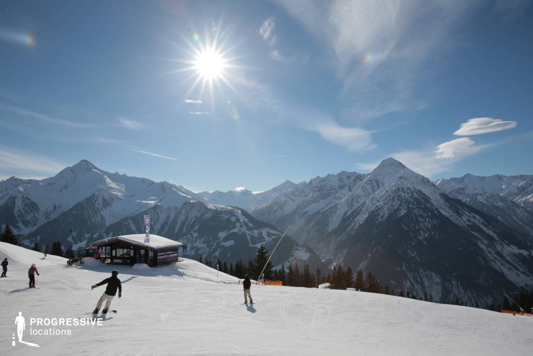 This wide mountain slope is positioned high above a deep alpine valley, offering sweeping views of rugged snow-covered peaks and forested ridges. In the midground stands a small, functional wooden instruction cabin marked for private ski lessons, subtly integrated into the gently sloping terrain. The foreground features a broad, groomed ski area with visible tracks and clear spatial orientation for downhill movement. The terrain transitions from open, snowy surfaces to dense pine forests along the valley edge, providing contrast in texture and color. The composition emphasizes the vastness of the landscape, with mountain silhouettes stretching across the horizon in soft, jagged lines, enhancing the visual depth. This setting is particularly suited for establishing expansive mountain scenes or sequences highlighting instructional and recreational infrastructure in a high-altitude context. Its combination of natural beauty and practical functionality supports a broad range of alpine production needs.
