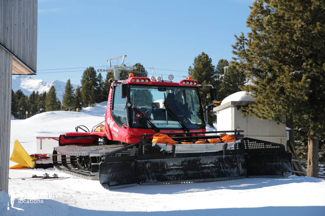 This image captures a heavy-duty snowcat vehicle parked at a high-altitude ski resort service area. The vehicle, painted in vivid red with black treads and a front-mounted grooming blade, is equipped for slope maintenance or emergency operations, as indicated by the attached rescue sled and utility accessories. The immediate environment includes compacted snow terrain and a backdrop of evergreen trees, with visible cable car infrastructure in the distance. The surrounding architecture features minimalist, functional structures, including a wooden-clad utility building and a concrete shelter. The snowcat’s industrial form, combined with the crisp grooming lines in the snow, introduces a technical and operational character to the natural alpine setting. This location is ideal for illustrating behind-the-scenes resort logistics, emergency preparedness, or mountain maintenance contexts. The contrast between the machine’s engineered form and the scenic mountain backdrop offers opportunities for dynamic compositions in documentary or operationally themed visual content.