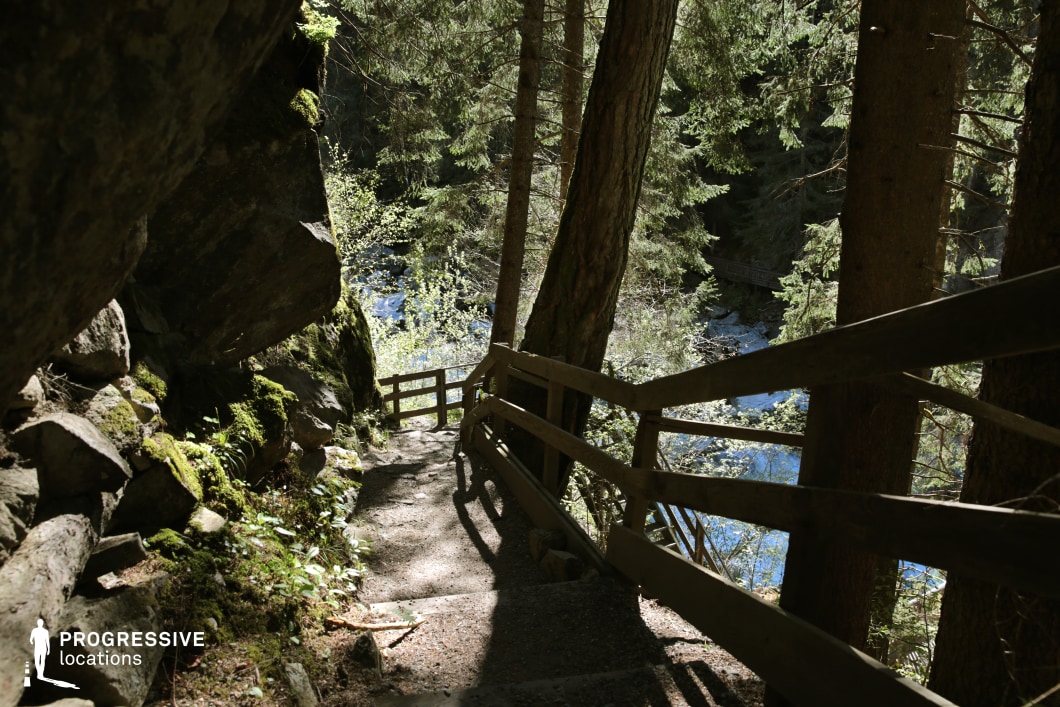 A narrow, shaded path winds steeply downward, bordered by a rustic wooden railing and carved into a rocky slope. Towering trees frame the descent, while massive moss-covered boulders hug one side and a glimpse of a shimmering river emerges through the foliage below. The interplay of light and shadow creates a layered, moody atmosphere ideal for suspense or introspective moments. This location is excellent for filming scenes in thriller, fantasy, or adventure genres—especially sequences involving escape, solitude, or a dramatic journey into the unknown.