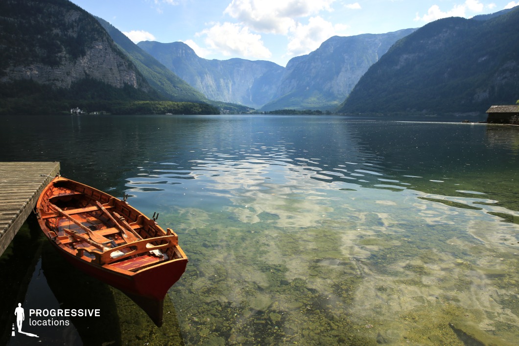 This tranquil lakeside setting features crystal-clear, shallow water transitioning into deep blues, bordered by towering cliffs and forested slopes. The red wooden rowboat at the weathered dock adds a rustic and timeless element to the composition. With its stillness, symmetry, and natural clarity, the scene is ideal for emotional or introspective moments in dramas, romance, or fantasy genres—especially scenes involving departures, reunions, or solitary reflection framed by majestic alpine serenity.