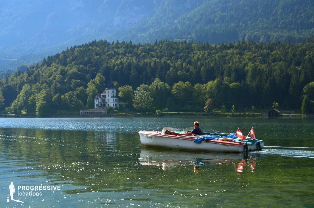 This scenic mountain lake is bordered by dense pine forests and overlooked by a striking white mansion with a turreted roof nestled at the tree line. The crystal-clear water reflects the greenery and distant hills, creating a serene and dreamlike atmosphere. This location is ideal for romance, mystery, or period dramas, particularly for scenes involving lakeside retreats, quiet introspection, or unfolding secrets in an elegant, secluded setting.