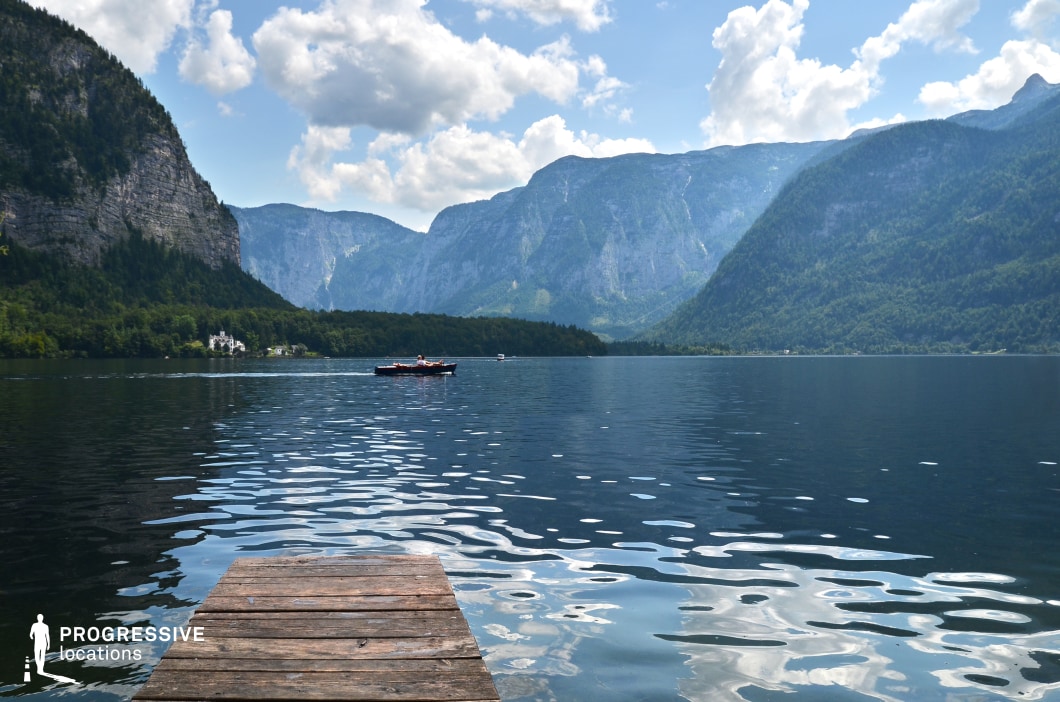 A simple wooden dock extends into a vast, tranquil mountain lake surrounded by towering, forested cliffs and dramatic rocky slopes. The stillness of the water mirrors the rugged landscape, while the distant boat and open horizon create a strong sense of depth and scale. The dock acts as a natural foreground anchor, inviting characters into the scene. This location is perfect for romantic, introspective, or adventurous moments in dramas, coming-of-age stories, or fantasy narratives where nature plays a central emotional or symbolic role.