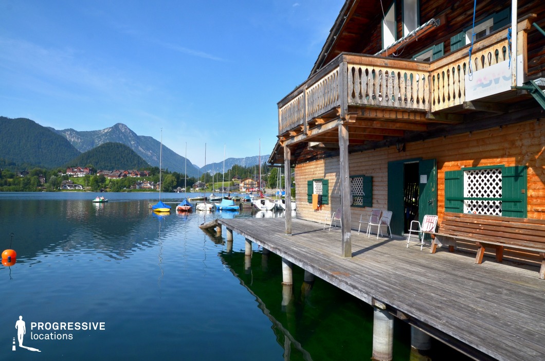 This inviting lakeside setting features a charming wooden boathouse with a broad dock extending over calm, clear waters, set against a backdrop of forested mountains and scattered sailboats. The combination of traditional architecture, tranquil marina, and scenic alpine surroundings evokes a peaceful and leisurely atmosphere. This location is perfect for romantic films, summer holiday stories, or character-driven dramas, especially for scenes involving quiet conversations, departures by boat, or serene moments of reflection near nature and water.