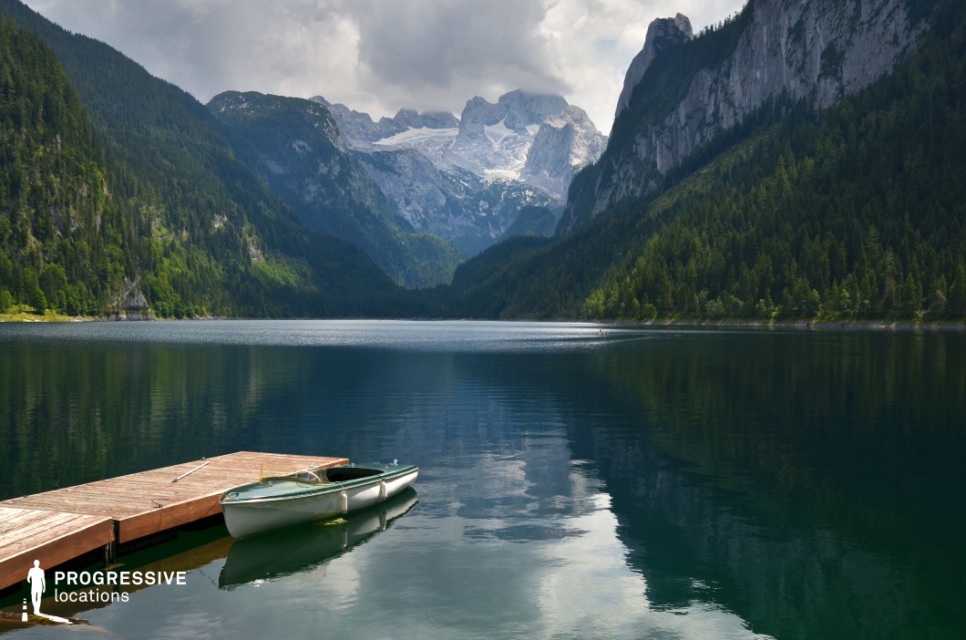 This breathtaking alpine lake is encircled by steep, forested mountains and capped by a dramatic view of snow-covered peaks in the distance. The calm, reflective water, paired with a simple dock and a moored boat, creates a striking yet peaceful composition. The strong sense of scale and isolation lends itself to emotionally charged or contemplative scenes in dramas, nature films, or fantasy epics—ideal for establishing shots, moments of solitude, or pivotal encounters framed by overwhelming natural beauty.