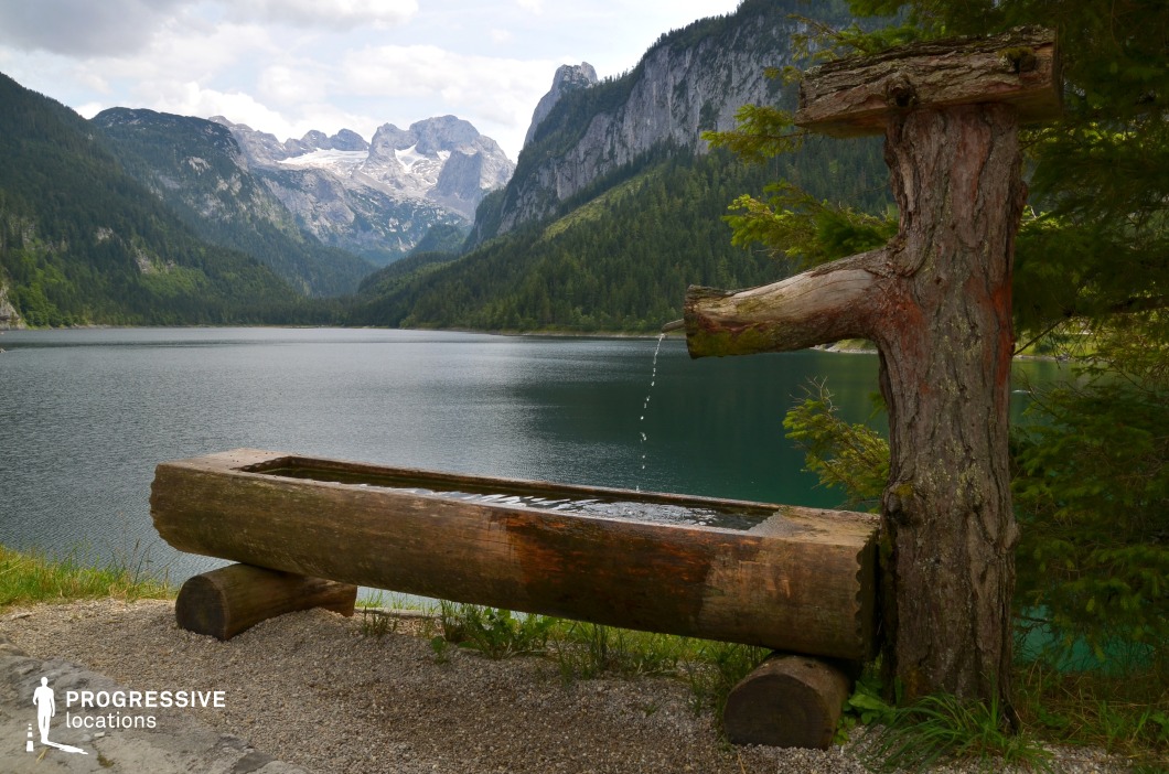 This tranquil mountain lake is framed by steep, forested slopes and towering, snow-capped peaks in the distance. In the foreground, a rustic wooden fountain crafted from logs trickles water into a trough, blending seamlessly into the natural environment. This location is ideal for filming serene and visually striking moments in dramas, nature documentaries, or fantasy films, especially scenes centered on solitude, spiritual reflection, or characters pausing during a long journey through pristine alpine wilderness.