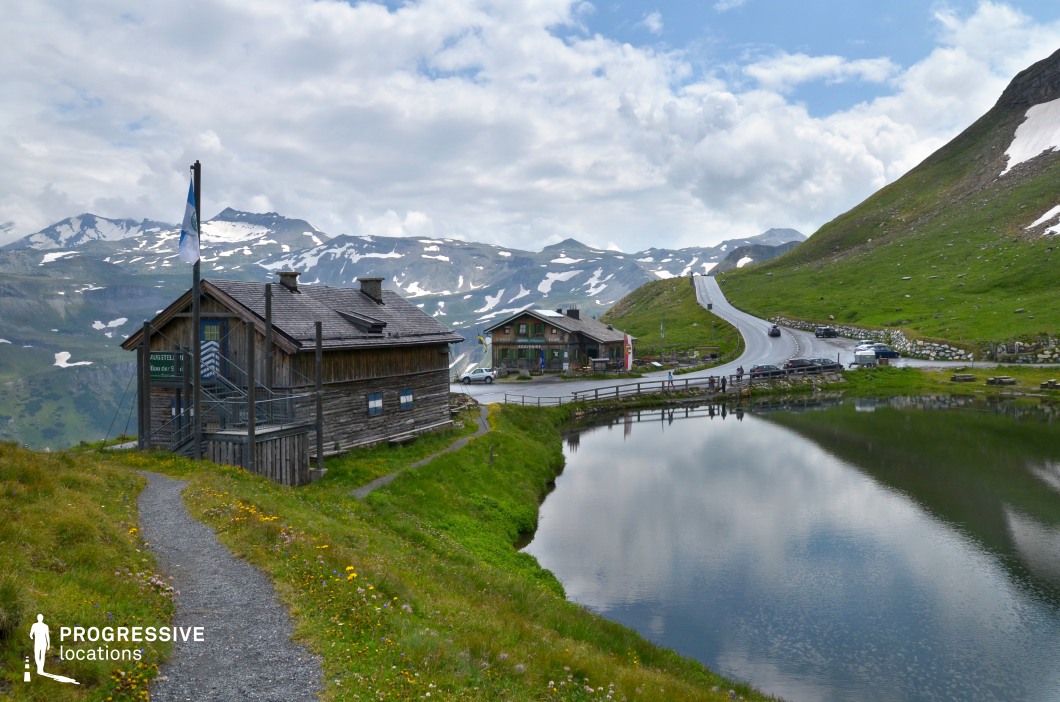This high-altitude location features two rustic wooden lodges nestled beside a small alpine lake, with a winding mountain road climbing into the snow-patched peaks beyond. The mix of natural beauty and human-made elements creates a balanced, scenic composition with a strong sense of place and elevation. Ideal for road movies, travel documentaries, or character-driven dramas, this setting supports scenes involving quiet retreats, transformative journeys, or pivotal rest stops in adventurous narratives.