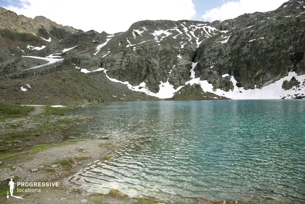 This striking alpine scene showcases a turquoise glacial lake bordered by rugged, snow-dusted rock formations and steep mountain walls. The lake’s still surface mirrors the stark surroundings, while patches of lingering snow emphasize the high-altitude, untouched environment. The shoreline blends gravel, moss, and stone, offering a natural foreground that transitions into the dramatic vertical backdrop. The absence of dense vegetation and the raw geological textures give the location a minimalist, elemental quality. Ideal for epic, survival, or introspective film scenes, this setting supports themes of isolation, endurance, or awe in a remote, high-altitude natural environment.