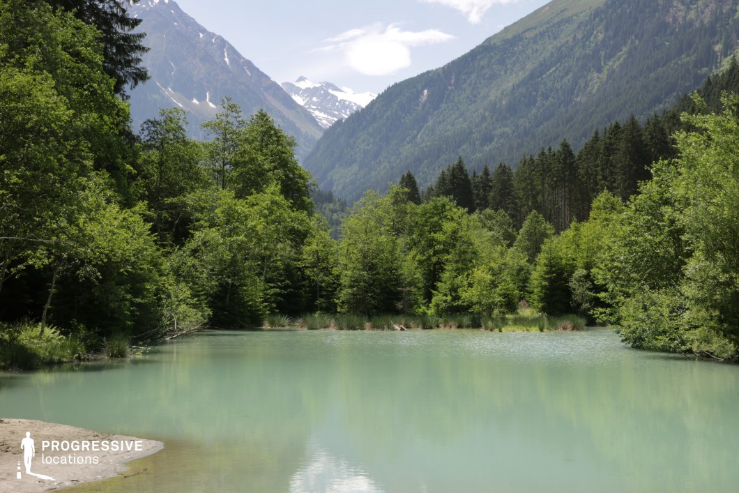 This tranquil alpine lake sits nestled in a lush green valley, surrounded by dense coniferous forest and flanked by towering, snow-dusted mountain slopes. The water has a distinctive pale turquoise hue, adding visual contrast to the dark green foliage and creating a serene, reflective surface. The soft shoreline blends naturally into the scene, enhancing the untouched, immersive quality of the location. This setting is ideal for films in the fantasy, adventure, or romantic genres, providing a scenic and isolated backdrop perfect for moments of reflection, natural wonder, or dramatic arrivals in a pristine wilderness.
