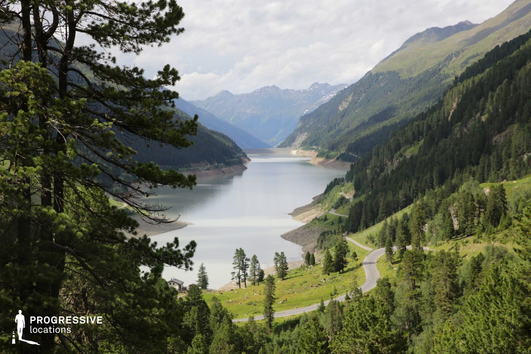 This expansive alpine vista showcases a long, serene reservoir nestled between steep, pine-covered slopes, with a winding mountain road tracing the hillside. The layered perspective, deep valley cut, and distant snowy peaks provide a sense of scale and isolation. The mixture of natural and infrastructural elements makes it suitable for scenic travel sequences, reflective solo journeys, or dramatic transitions in road movies, thrillers, or epic adventures.