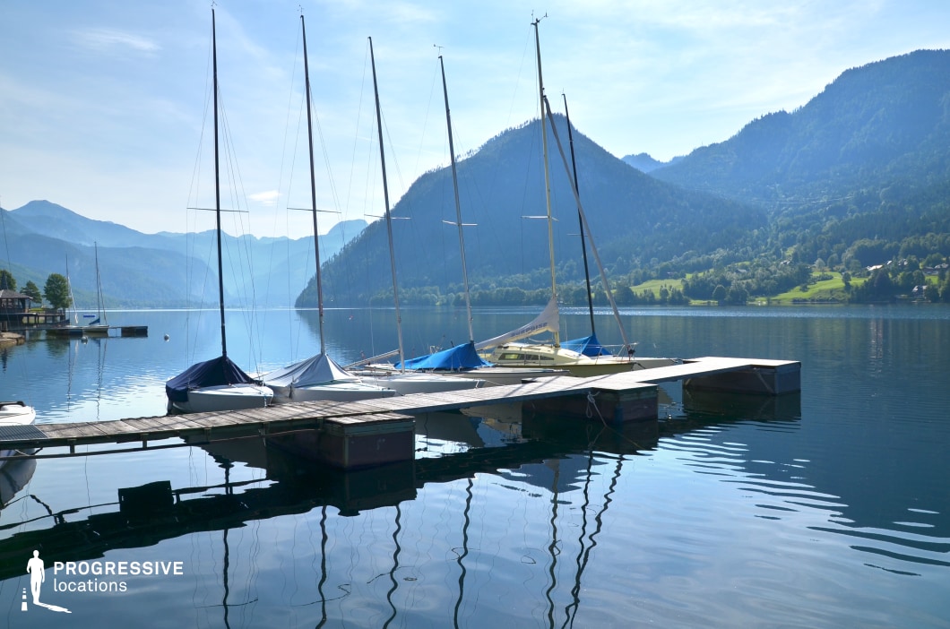 This elegant lakeside dock hosts a row of moored sailboats, their masts mirrored perfectly in the still water, set against a sweeping panorama of layered alpine hills and distant ridgelines. The blend of precision and natural beauty evokes calm, leisure, and escape. This location is perfect for refined or emotionally subtle scenes in dramas, romantic films, or travel-focused narratives—especially moments of arrival, departure, quiet reflection, or bonding set in an upscale or serene lakeside environment.