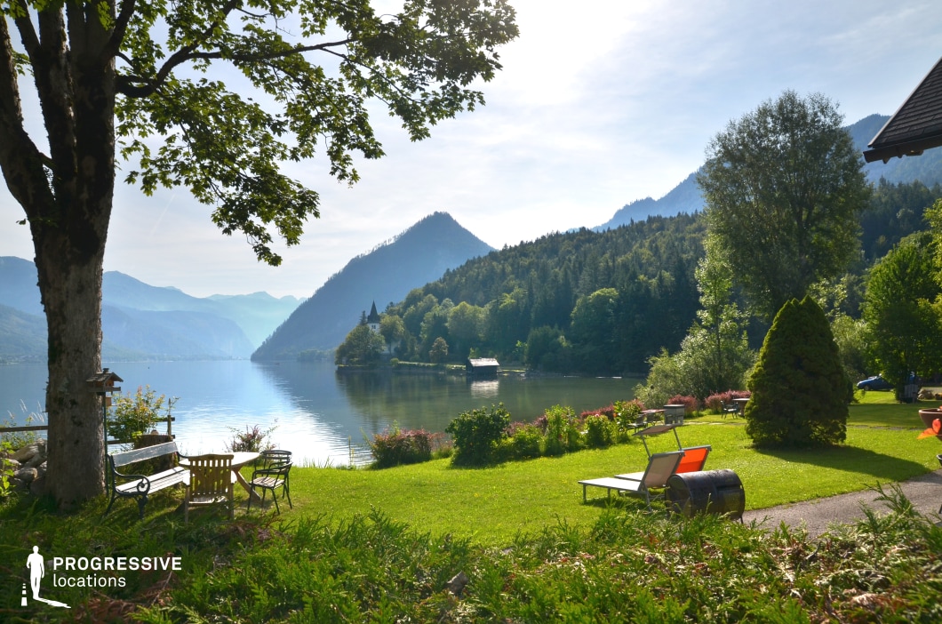 This picturesque location features a manicured lakeside garden framed by lush greenery, looking out onto a calm alpine lake surrounded by forested hills and distant peaks. The composition offers layered depth, from the shaded foreground to the sunlit mountain slopes, creating a serene and harmonious visual balance. Ideal for romantic or reflective scenes, upscale retreats, or lakeside conversations, this setting suits dramas, romantic films, travel features, or character-centered stories set in tranquil or luxurious environments.