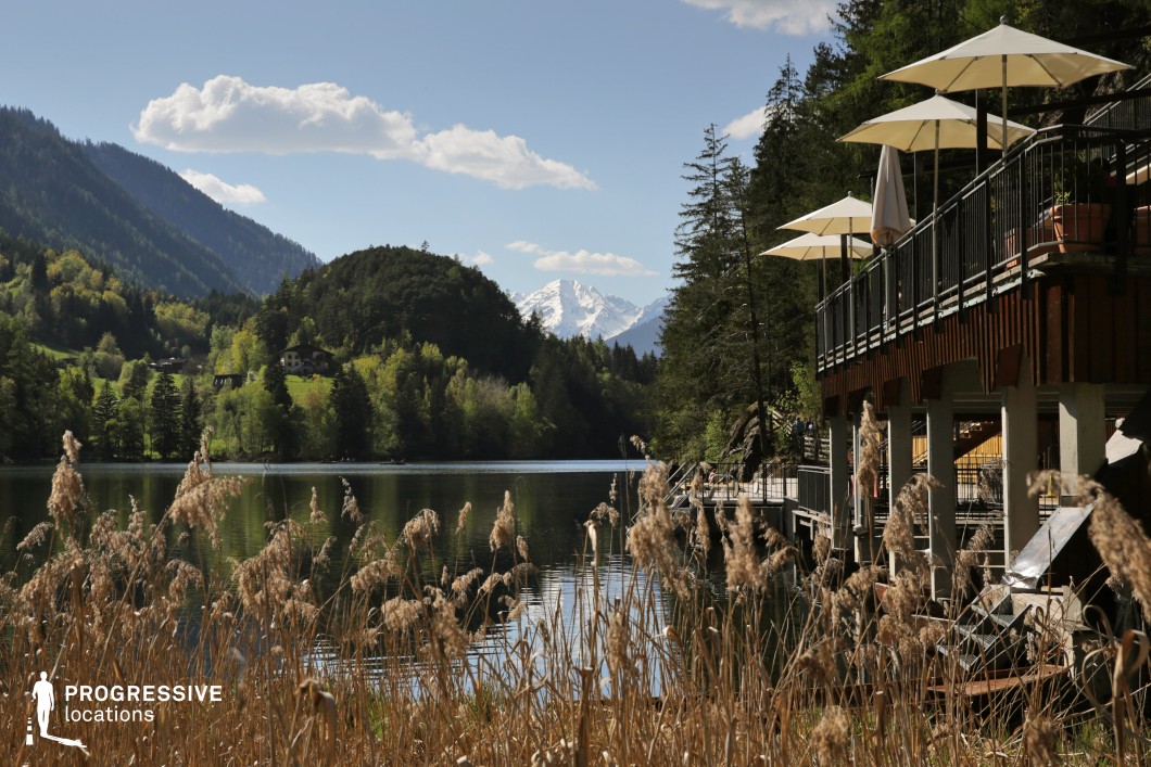 This scenic lakeside location blends natural elegance with leisure, featuring a tranquil body of water flanked by forested hills and a snow-capped peak in the distance. A stylish wooden terrace with umbrellas adds a touch of refinement, suggesting a serene retreat or upscale setting. The framing layers foreground reeds, midground architecture, and dramatic background mountains, making it ideal for romantic or introspective scenes, high-end travel features, or character-driven narratives set in exclusive or peaceful alpine environments.