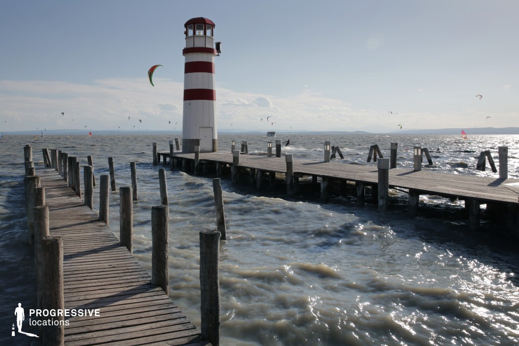This striking waterfront location features a bold red-and-white striped lighthouse at the end of a wooden pier, surrounded by choppy waters and distant kite surfers. The rhythmic structure of the pier, paired with the motion of the wind and water, creates a dynamic and energetic visual tension. The open horizon and nautical iconography make this setting ideal for adventure films, emotional climaxes, or introspective sequences—especially those involving departures, reunions, or scenes that demand a powerful coastal backdrop.