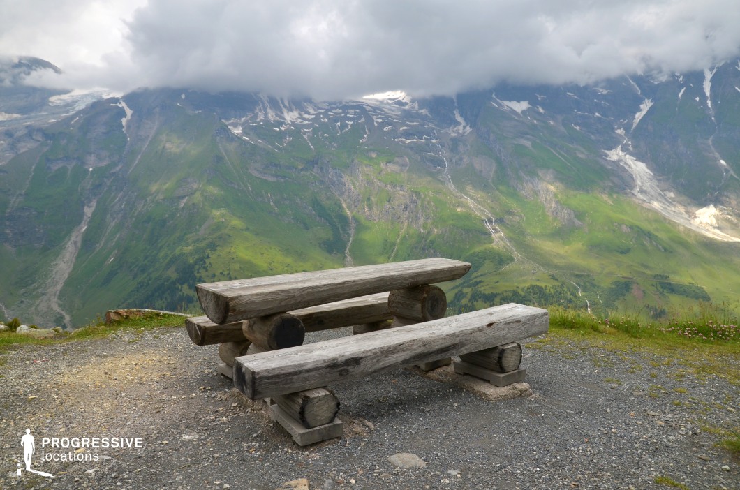 This mountaintop location features a rustic log bench and table set at the edge of a dramatic cliff, facing an expansive panorama of snow-streaked peaks and steep green valleys. The raw simplicity of the seating contrasts with the grandeur of the natural backdrop, offering a quiet focal point in an overwhelming landscape. Ideal for character-driven scenes of reflection, emotional turning points, or pivotal conversations, this spot suits dramas, survival films, or epics that draw power from isolation, scale, and the sublime beauty of the high mountains.