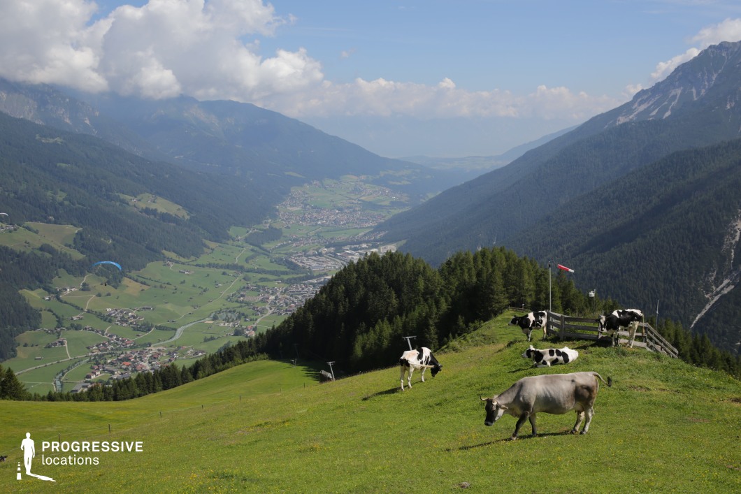 This expansive alpine meadow overlooks a deep, forested valley with winding roads, scattered villages, and layered mountain ridges fading into the distance. The lush green hillside in the foreground offers open space and natural elevation, with a fence and windsock suggesting a high-altitude vantage point. The background reveals a detailed rural settlement layout, framed by forested slopes and rugged peaks. This panoramic location is highly suitable for establishing shots, travel sequences, or pastoral scenes in adventure, nature documentary, or period films that require a sweeping, tranquil yet elevated mountain environment.